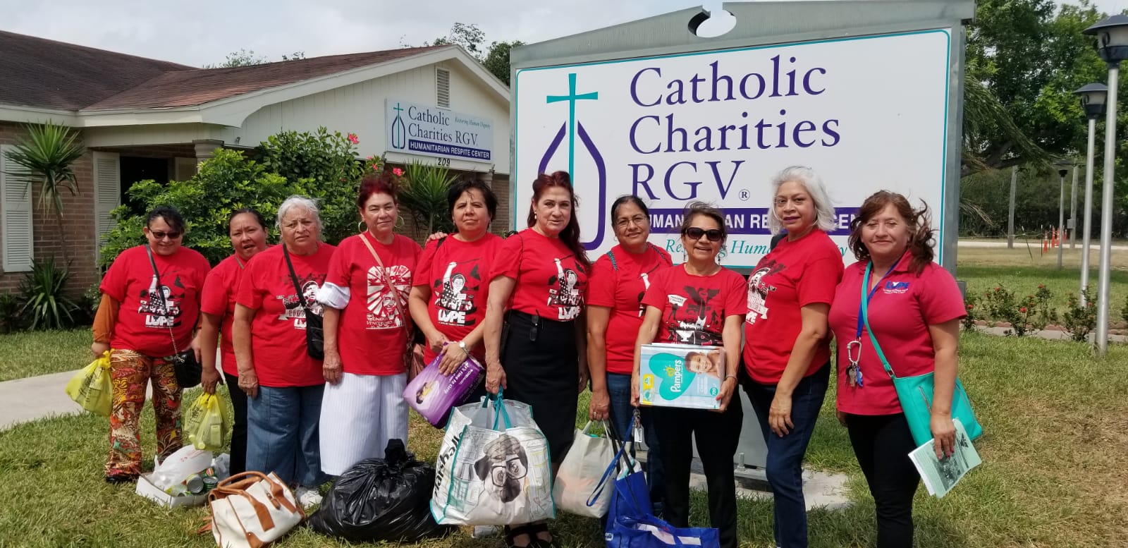 Group of women LUPE members dressed in red shirts hold supply donations in bags and boxes in front of sign that reads "Catholic Charities RGV"